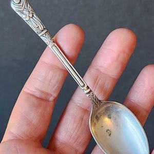 Puede incluir: Una cuchara de plata con un mango adornado con un diseño floral. La cuchara tiene un cuenco redondeado y mide aproximadamente 10 cm de largo.