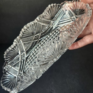 Könnte beinhalten: Eine klare Glasservierplatte mit einem dekorativen Schliffmuster. Die Platte ist oval geformt und hat einen gewellten Rand.