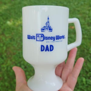 May include: White ceramic mug with a blue Walt Disney World logo and the word "DAD" printed on it. The logo features the Cinderella Castle and Mickey Mouse head.
