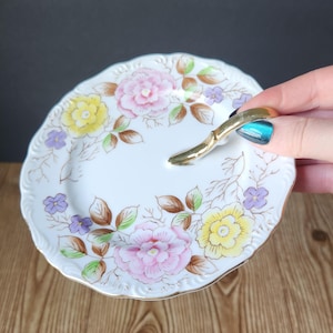 May include: A white porcelain serving plate with a gold handle and floral design. The plate features pink, yellow, and purple flowers with brown leaves.