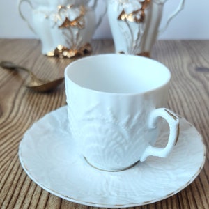 May include: A white porcelain teacup and saucer set with a delicate floral design and gold accents. The cup and saucer have a raised, textured surface.