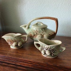 May include: A vintage tea set featuring a teapot and two creamers. The set is light green with brown accents and a leaf and fruit design. The teapot has a curved handle and a lid. The creamers have handles and a spout.
