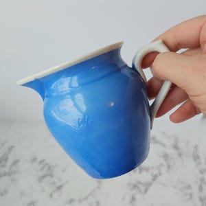Może przedstawiać: Mały, niebieski ceramiczny dzbanek z białym rantem i uchwytem. Dzbanek ma błyszczące wykończenie i zakrzywiony dzióbek. Uchwyt jest w kolorze jasnoszarym. Dzbanek jest trzymany w dłoni.