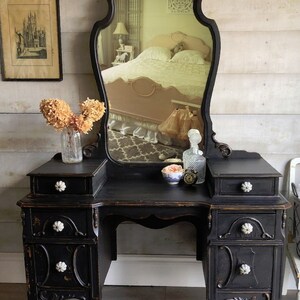 Puede incluir: Un tocador negro con un gran espejo adornado y cuatro cajones. El tocador tiene pomos de porcelana blanca y se asienta sobre cuatro patas torneadas. El espejo tiene un borde biselado y una parte superior decorativa.