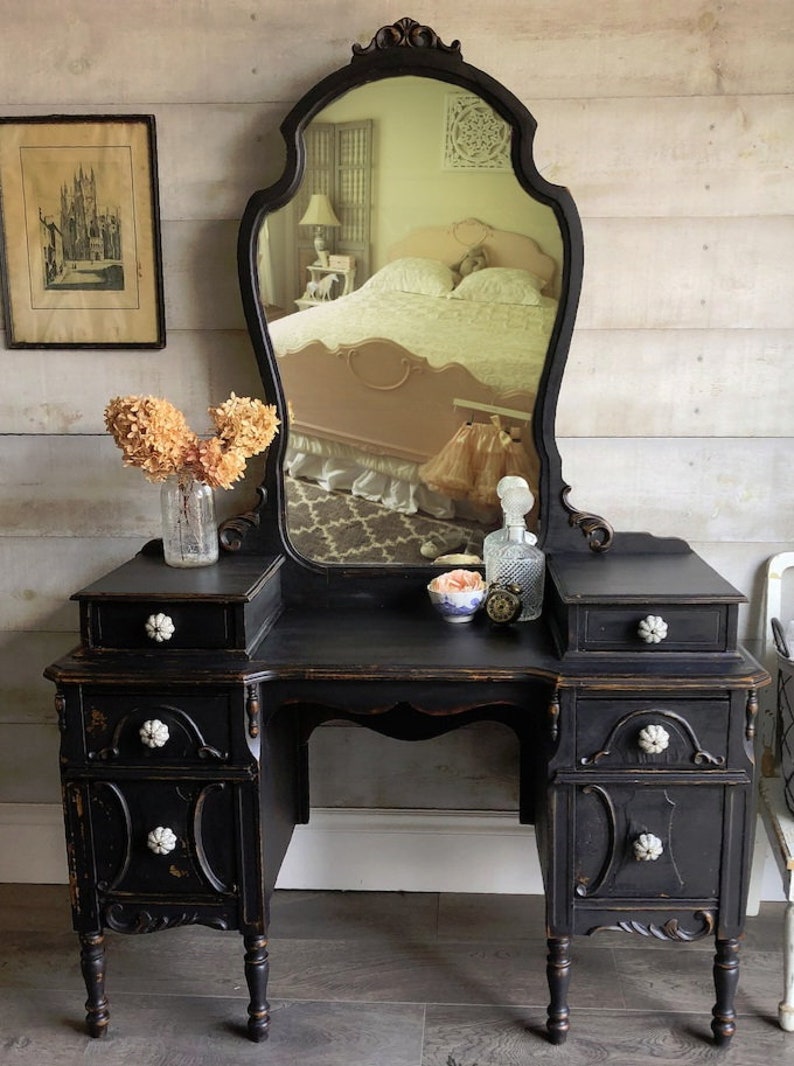 Puede incluir: Un tocador negro con un gran espejo adornado y cuatro cajones. El tocador tiene pomos de porcelana blanca y se asienta sobre cuatro patas torneadas. El espejo tiene un borde biselado y una parte superior decorativa.