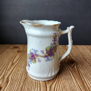 May include: A white porcelain pitcher with a gold rim and a floral design featuring purple and blue flowers. The pitcher has a handle and a scalloped rim.