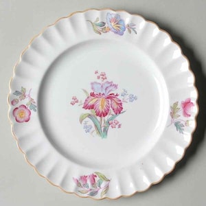 May include: White ceramic plate with a scalloped edge and a gold rim. The plate features a central floral design with pink, purple, and yellow flowers, and smaller floral accents around the rim. A decorative piece for serving.