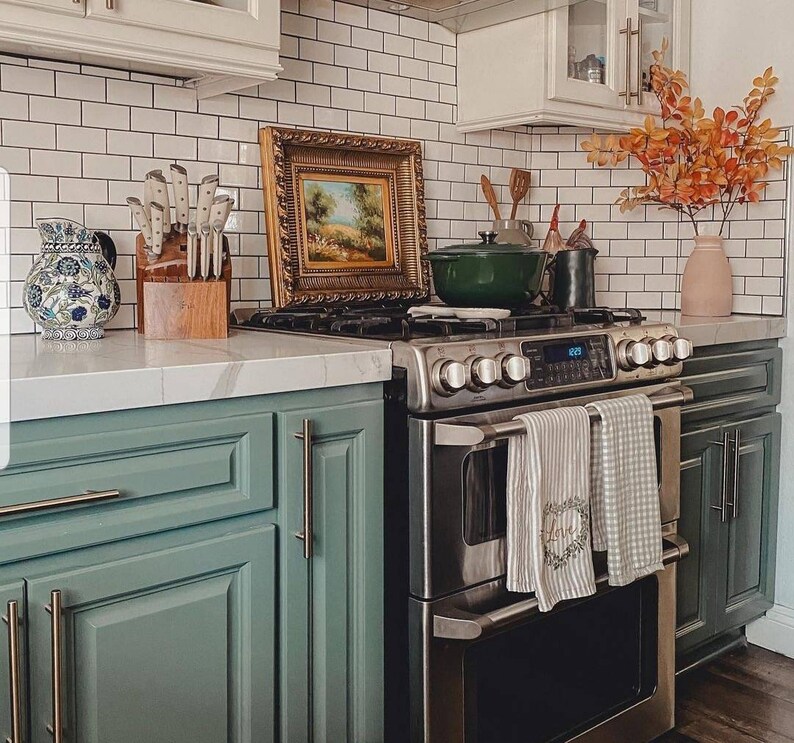 May include: A stainless steel double oven with a black glass cooktop. The oven has a digital display and is set into a green kitchen cabinet. There are two white towels hanging from the oven door.