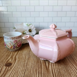 Puede incluir: Una tetera de cerámica rosa claro con tapa cuadrada y asa a juego, colocada junto a una taza de té blanca con un colorido estampado floral de rosas y borde dorado, sobre un fondo de azulejos blancos y una superficie de madera.