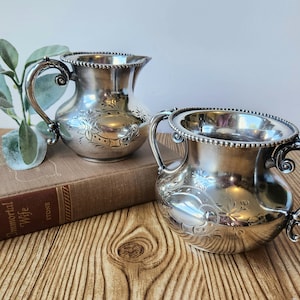 Könnte beinhalten: Ein silbernes Sahne- und Zuckerdosen-Set. Die Sahnekännchen hat einen Ausguss und einen Griff, die Zuckerdose hat zwei Griffe. Beide Teile weisen florale Gravuren und Perlenränder auf. Das Set steht auf einer Holzoberfläche.
