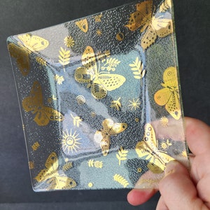 以下が含まれることがあります： 金色の縁取りと蝶と渦巻き模様が施された正方形のガラス製サービングディッシュ。ディッシュは金色のメタリックペイントで装飾されています。