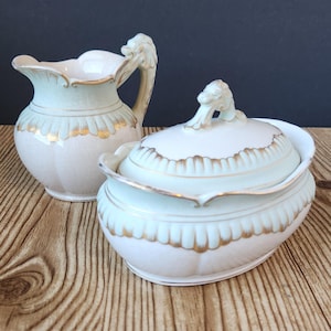 May include: A vintage porcelain creamer and sugar bowl set with a light green and white design and gold accents. The lid and handle of each piece feature a decorative lion head.