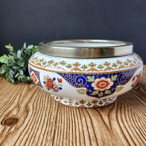 Puede incluir: Un cuenco de cerámica blanca con un borde plateado y un patrón floral colorido. El cuenco tiene un diseño floral azul, dorado y rojo sobre un fondo blanco.