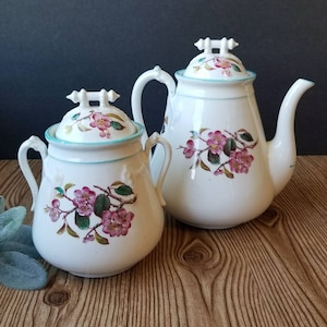Puede incluir: Una tetera y un azucarero de porcelana blanca con diseños florales. La tetera tiene una asa y un pico, mientras que el azucarero tiene dos asas. Ambas piezas presentan un delicado patrón floral con acentos rosas y verdes.