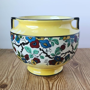 Può includere: Un vaso in ceramica giallo con manici neri e un motivo floreale in tonalità di blu, rosso, verde e nero. Il vaso ha un corpo rotondo e un bordo svasato.