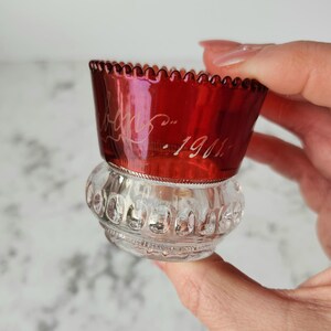 Può includere: Un piccolo vaso di vetro trasparente con un top in vetro rosso. Il top ha un bordo smerlato ed è inciso con la data "Dic. 1911".