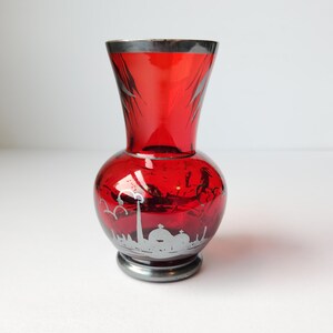 May include: A red glass vase with a silver rim and base. The vase has a bulbous body and a flared top. The body features a silver cityscape design. The vase is approximately 6 inches tall.