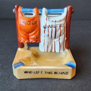 May include: A ceramic ashtray shaped like a clothesline with two hanging items. One item is labeled "Matches" and the other is labeled "Cigarettes". The ashtray has a blue and white trim and the words "Who left this behind" are printed on the bottom.