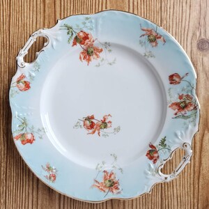 Puede incluir: Una fuente redonda y antigua con borde celeste y centro blanco. La fuente presenta un diseño floral de amapolas rojas y follaje verde. Los bordes y asas están adornados con detalles dorados, añadiendo un toque de elegancia a esta pieza vintage.
