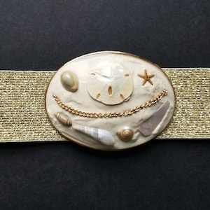May include: A white oval belt buckle with a gold chain and various seashells, including a sand dollar, starfish, and a conch shell. The buckle is attached to a gold glitter elastic belt.