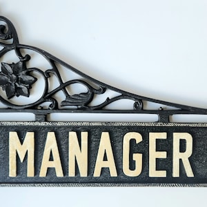 Puede incluir: Un letrero de hierro fundido negro con un soporte decorativo de volutas. El letrero dice "MANAGER" en letras blancas sobre fondo negro.