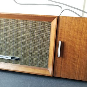 Op de afbeelding: Een rechthoekige Panasonic 2-way system luidspreker met een houten frame en een textuurrijk olijfgroen stoffen rooster. Het apparaat heeft een klein rood indicatielampje in de linkerbovenhoek en een massief houten paneel aan de rechterkant met een verticale handgreep.