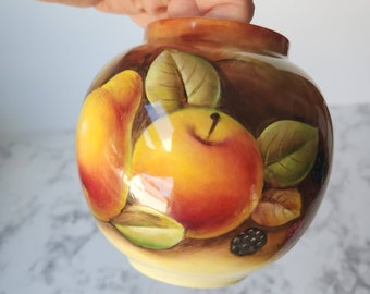 May include: A small, round ceramic vase with a hand-painted fruit design. The vase features a pear, apple, and leaves in shades of yellow, orange, and green against a brown background. The vase is held by a hand.