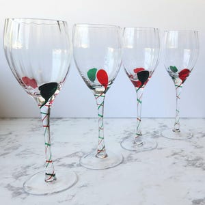 May include: Four clear wine glasses with a decorative design. Each glass has a red, green, and black painted design at the base of the bowl. The stems are clear with red and green stripes. The glasses are on a white surface.