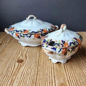 May include: Two white porcelain serving dishes with lids. The dishes feature a floral pattern with orange and blue accents. The lids have a handle on top and the smaller dish has a spoon opening. The dishes are on a wooden surface.
