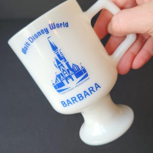 May include: White ceramic mug with a blue and white image of Cinderella's Castle from Walt Disney World. The mug has a pedestal base and the text "Walt Disney World" and "BARBARA" printed on it.