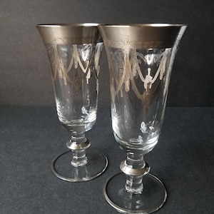 May include: Two clear champagne flutes with silver rims and decorative etched patterns. The flutes have a classic design with a round base and a long stem. The silver rim and etched details add a touch of elegance to the glassware.