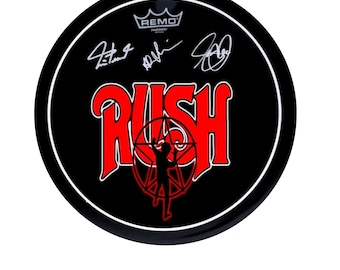 May include: A black and red drumhead with the word "RUSH" in red letters and a stylized image of a person in a circle. The drumhead is signed by the band members: Geddy Lee, Alex Lifeson, and Neil Peart. The drumhead is a Remo Pinstripe.