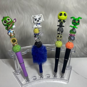 May include: Four decorative pens with character-themed toppers. The pens feature a purple pen with a green frog, a blue pen with a Dalmatian, a black pen with Jack Skellington, and a black pen with a green alien. Each pen has decorative beads.