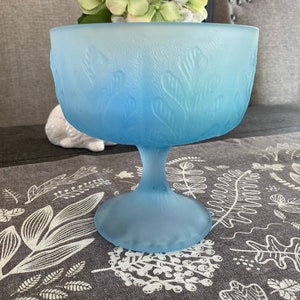 May include: A frosted blue glass pedestal bowl with a floral design. The bowl is sitting on a gray and white patterned tablecloth.