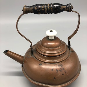 May include: A vintage copper tea kettle with a black wooden handle and a white porcelain knob on the lid.