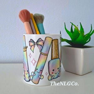 May include: A white ceramic mug used as a makeup brush holder, featuring a colourful rainbow gradient print of cartoon beauty tools with faces, a bow, and stars. The mug contains three makeup brushes with peach, yellow, and teal handles.