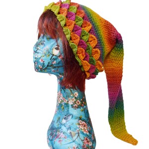 May include: A vibrant, handmade crochet hat with a long, pointed tail. The hat showcases a rainbow gradient of colours, including green, orange, pink, and yellow. The hat is displayed on a blue mannequin head with a floral pattern.