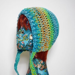 May include: A handmade crochet hat in shades of blue, green, and orange. The hat has ear flaps and a textured stitch pattern. The hat is displayed on a mannequin head with a floral pattern.