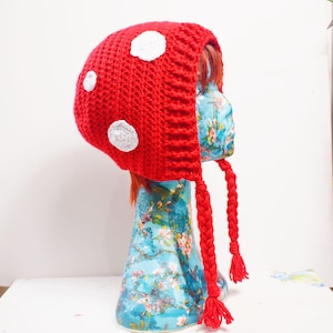 May include: A red crocheted hat with white polka dots, resembling a mushroom. The hat has long, braided tassels and is displayed on a floral patterned mannequin head. This whimsical hat is perfect for costume parties or everyday wear.