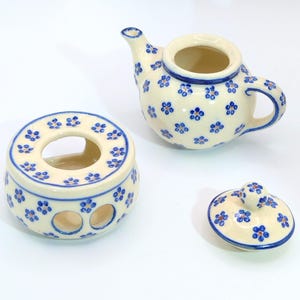 May include: A cream-colored ceramic teapot, warmer, and lid set. The pieces are decorated with blue floral patterns and blue trim. The teapot has a curved handle and spout. The warmer has three openings. The lid is round.