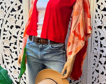 May include: A red cropped jacket with a pink collar and floral patterned sleeves. The jacket is worn over a white tank top and blue jeans. A brown hat is held in the hand.