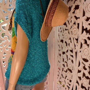 May include: A turquoise knitted top with a rainbow pattern and short sleeves. The top is worn with denim shorts and a brown woven hat.
