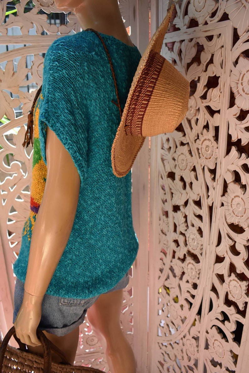 May include: A turquoise knitted top with a rainbow pattern and short sleeves. The top is worn with denim shorts and a brown woven hat.
