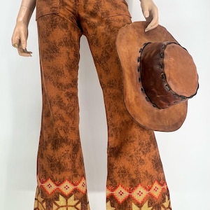 May include: Brown flared pants with a textured pattern and decorative embroidery at the hem. A brown leather hat is held next to the pants. The pants have two front pockets and a high waist. The pants are a vintage style.
