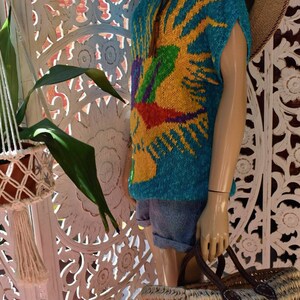 May include: A turquoise knitted sweater with a colorful sun design. The sweater has short sleeves and a relaxed fit. The sweater is displayed on a mannequin with a straw hat and a woven bag.
