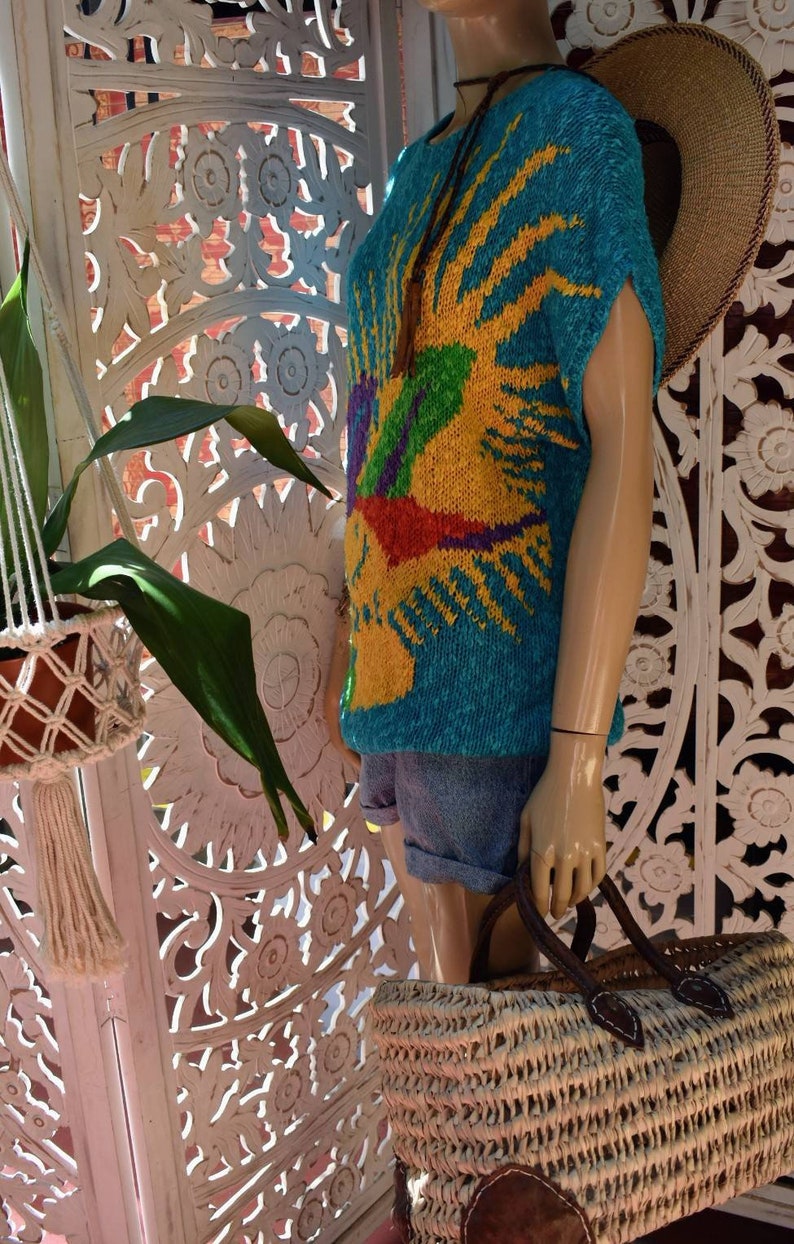 May include: A turquoise knitted sweater with a colorful sun design. The sweater has short sleeves and a relaxed fit. The sweater is displayed on a mannequin with a straw hat and a woven bag.