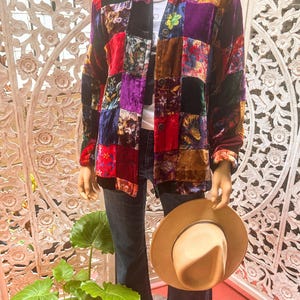 May include: A multi-colored patchwork velvet shirt with a button-up front and long sleeves. The shirt is made of different fabrics and colors, including red, purple, brown, and black. The shirt is being modeled on a mannequin.