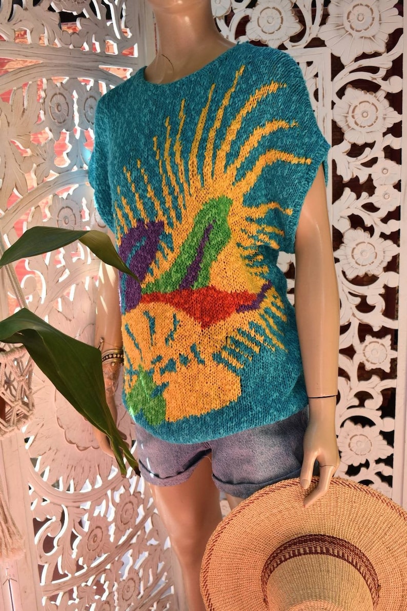 May include: A teal blue knit sweater with a colorful abstract design. The design features a yellow sunburst with green, purple, and red accents.
