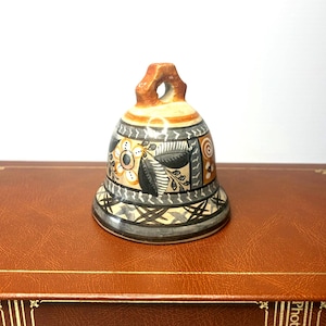 May include: A ceramic bell featuring a hand-painted design with black, white, and orange floral and geometric patterns. The bell has a rounded top handle and a flared base, with intricate leaf motifs and cross-hatch details encircling the exterior surface.