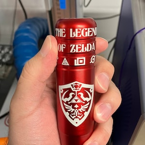 May include: A red metal tube with the words "THE LEGEND OF ZELDA" and a white stylized image of the Triforce and the Hylian Crest.